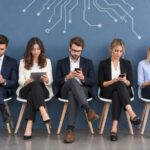 Pet mladih profesionalaca sedi u redu i koristi mobilne telefone i tablete u modernom poslovnom prostoru